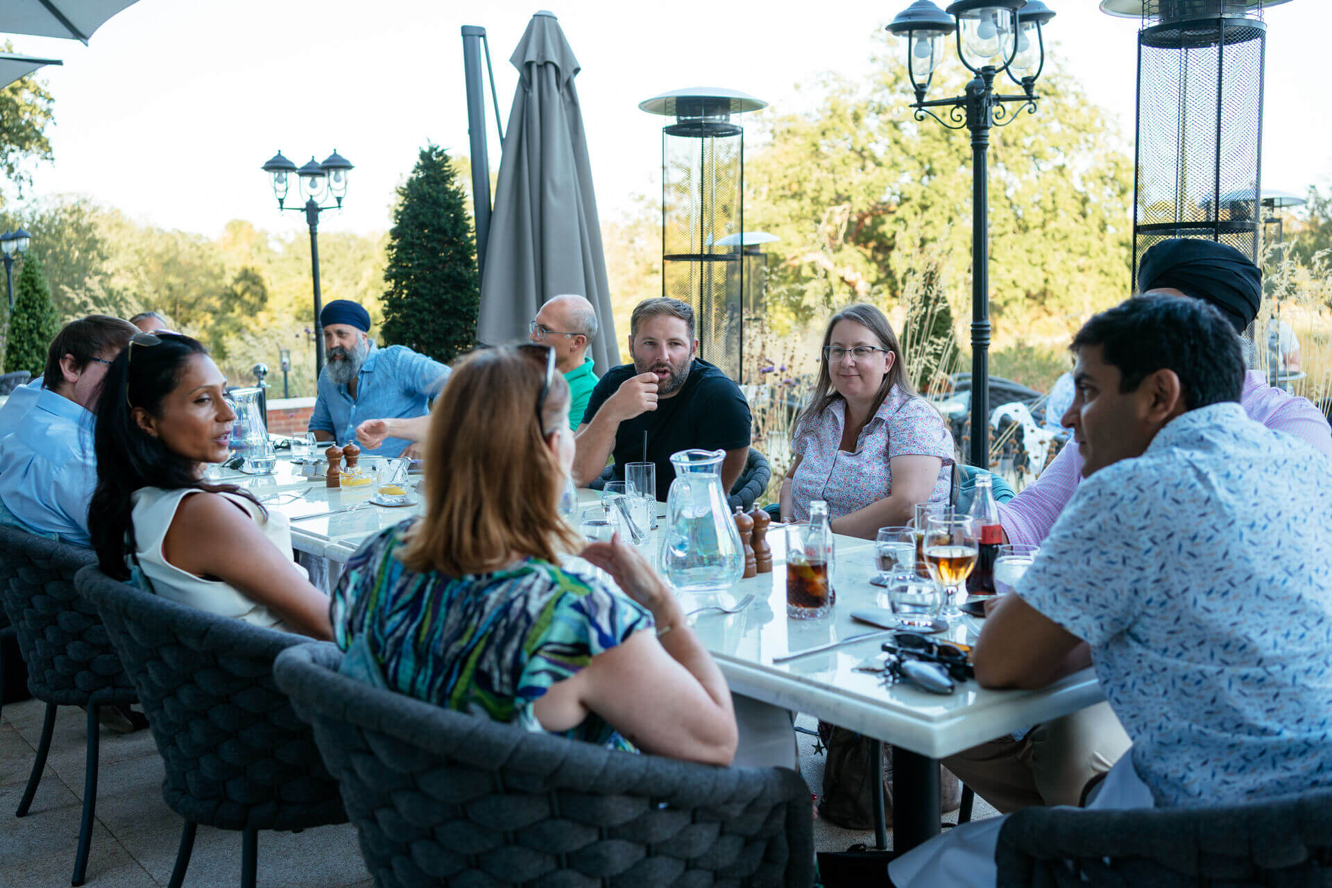 A group of attendees talk over drinks and a meal around an outdoor dining table.