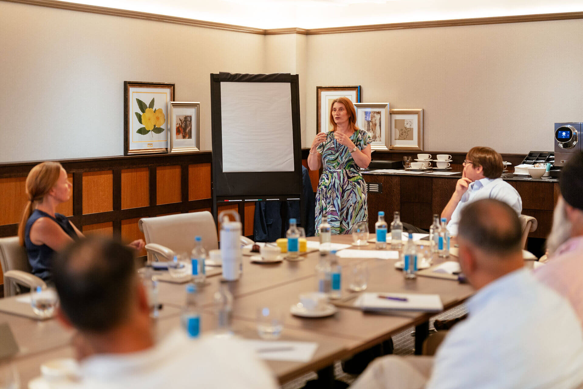 A woman leads a workshop beside a blank flip chart while attendees sit around a conference table.